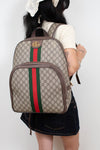 A person shown from the back wearing a beige Gucci backpack with the classic GG monogram pattern, brown leather trim, and the signature green and red web stripe down the center.