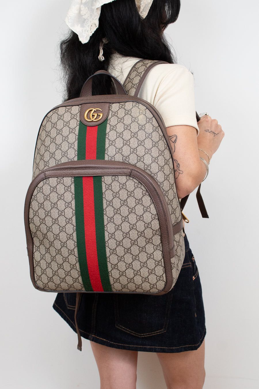 A person shown from the back wearing a beige Gucci backpack with the classic GG monogram pattern, brown leather trim, and the signature green and red web stripe down the center.