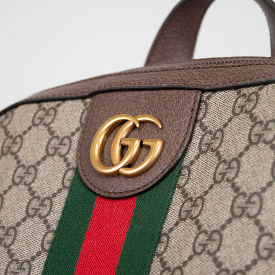 Close-up of a Gucci backpack featuring the brand's GG monogram coated canvas, brown leather trim, a green and red web stripe, and a gold-toned GG logo detail.