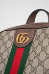 Close-up of a Gucci backpack featuring the brand's GG monogram coated canvas, brown leather trim, a green and red web stripe, and a gold-toned GG logo detail.