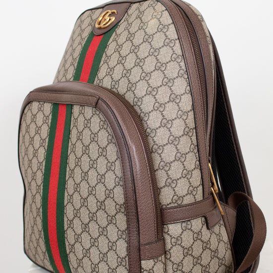 Gucci GG Supreme backpack in beige and brown monogram coated canvas with the signature green and red web stripe, brown leather trim, and gold-tone hardware, shown on a light background.