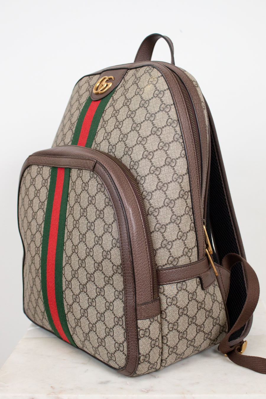 Gucci GG Supreme backpack in beige and brown monogram coated canvas with the signature green and red web stripe, brown leather trim, and gold-tone hardware, shown on a light background.