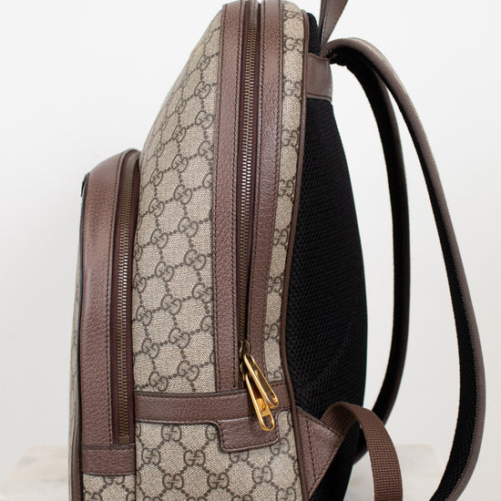 A Gucci backpack shown from the side, featuring the brand's signature GG Supreme coated canvas in beige and brown, brown leather trim, a black mesh back panel, and gold-tone hardware.