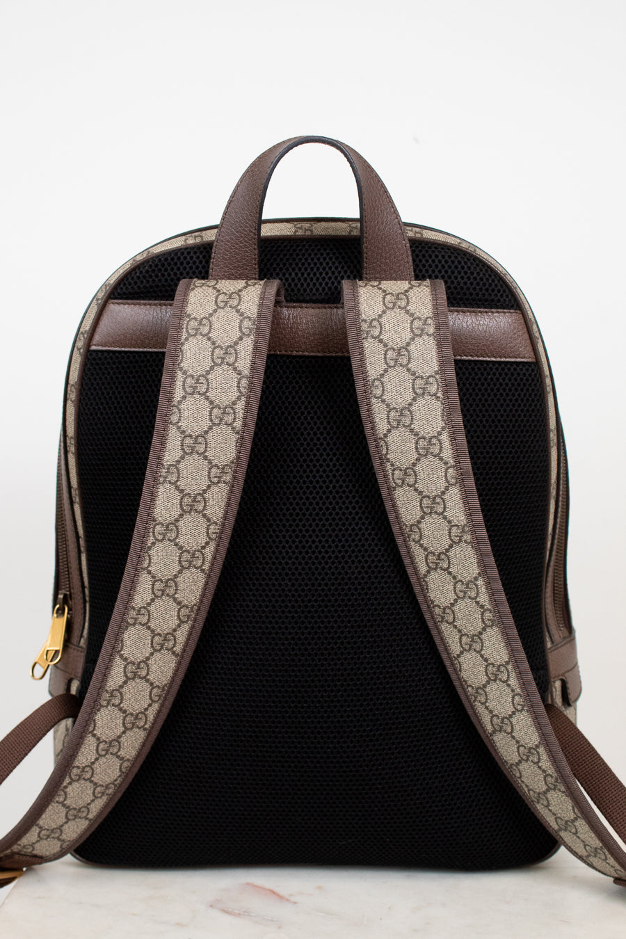 The back view of a Gucci backpack featuring a black mesh back panel, brown leather trim, and beige and brown GG monogram coated canvas shoulder straps.