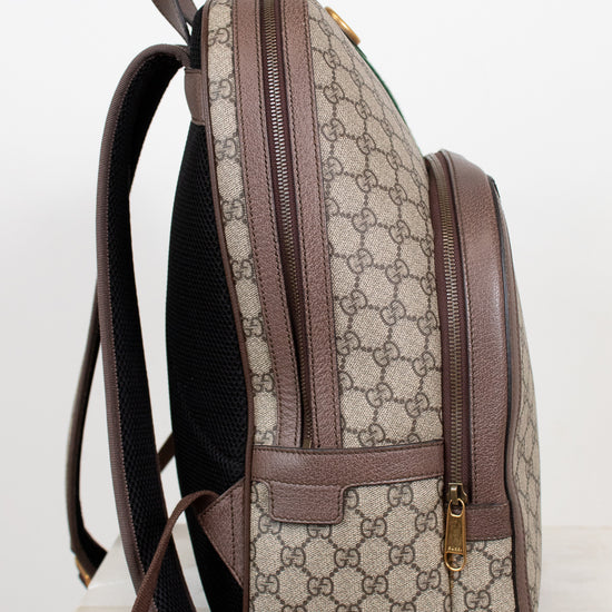 A Gucci backpack in beige GG Supreme coated canvas with brown leather trim, shown from a side profile view on a light marble surface against a white background.