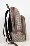 A Gucci backpack in beige GG Supreme coated canvas with brown leather trim, shown from a side profile view on a light marble surface against a white background.