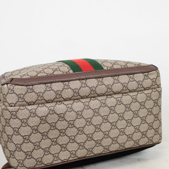 Bottom view of a Gucci backpack in beige GG Supreme coated canvas with brown leather trim and the signature green and red web stripe, resting on a light marble surface against a white background.