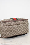 Bottom view of a Gucci backpack in beige GG Supreme coated canvas with brown leather trim and the signature green and red web stripe, resting on a light marble surface against a white background.