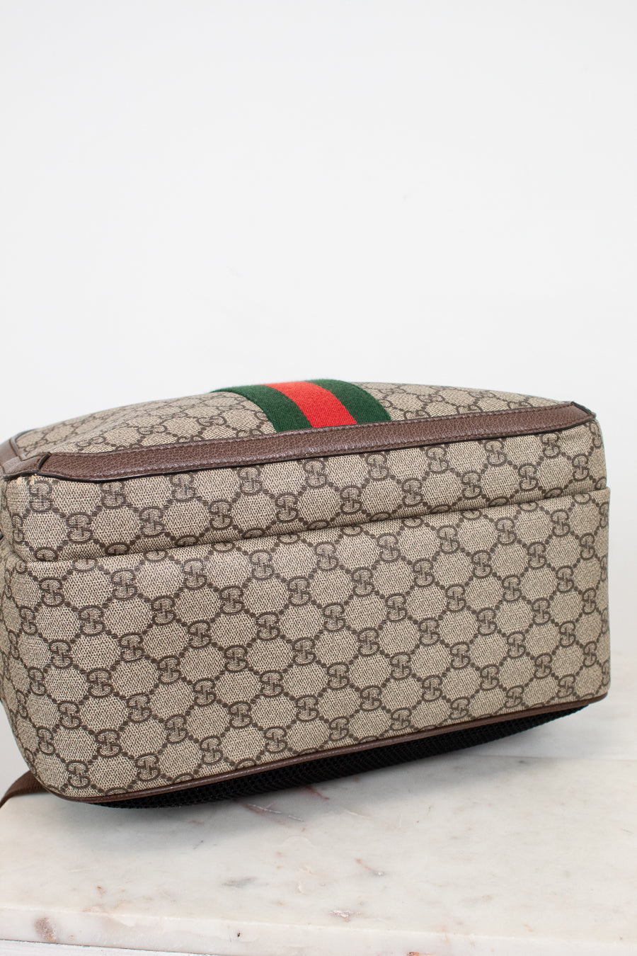 Bottom view of a Gucci backpack in beige GG Supreme coated canvas with brown leather trim and the signature green and red web stripe, resting on a light marble surface against a white background.