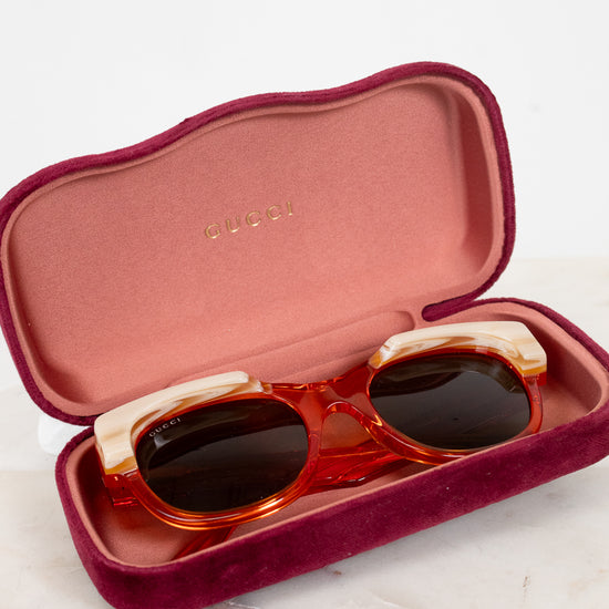 Gucci round sunglasses with translucent orange and cream frames and dark lenses, resting in an open burgundy velvet case with 'Gucci' visible on a light marble surface.