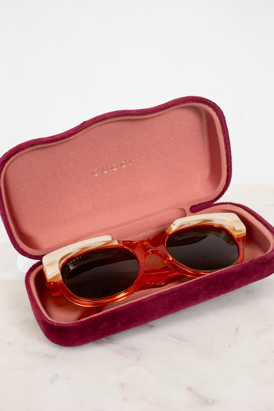 Gucci round sunglasses with translucent orange and cream frames and dark lenses, resting in an open burgundy velvet case with 'Gucci' visible on a light marble surface.