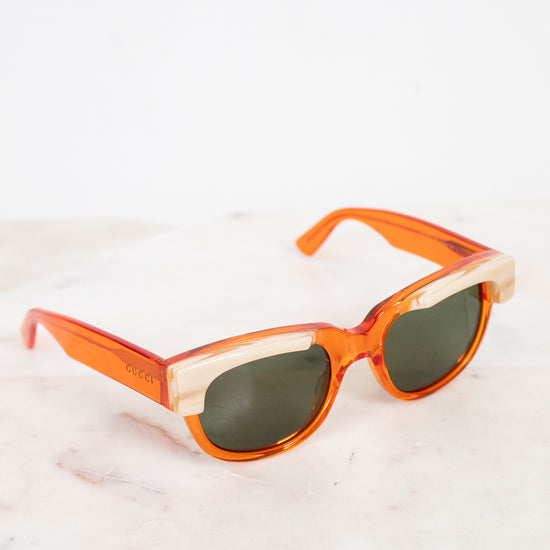 A pair of Gucci sunglasses with translucent orange acetate frames, cream marbled detail and dark lenses, resting on a white marble surface against a white background.