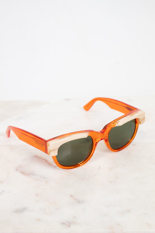 A pair of Gucci sunglasses with translucent orange acetate frames, cream marbled detail and dark lenses, resting on a white marble surface against a white background.