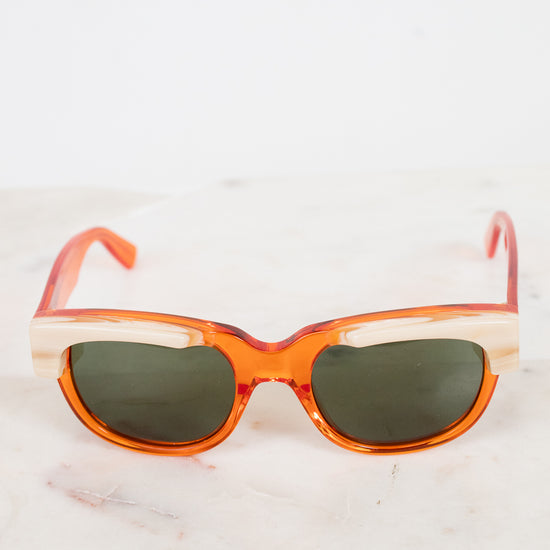 Orange acetate sunglasses with cream details and dark lenses resting on a white marble surface against a white background.