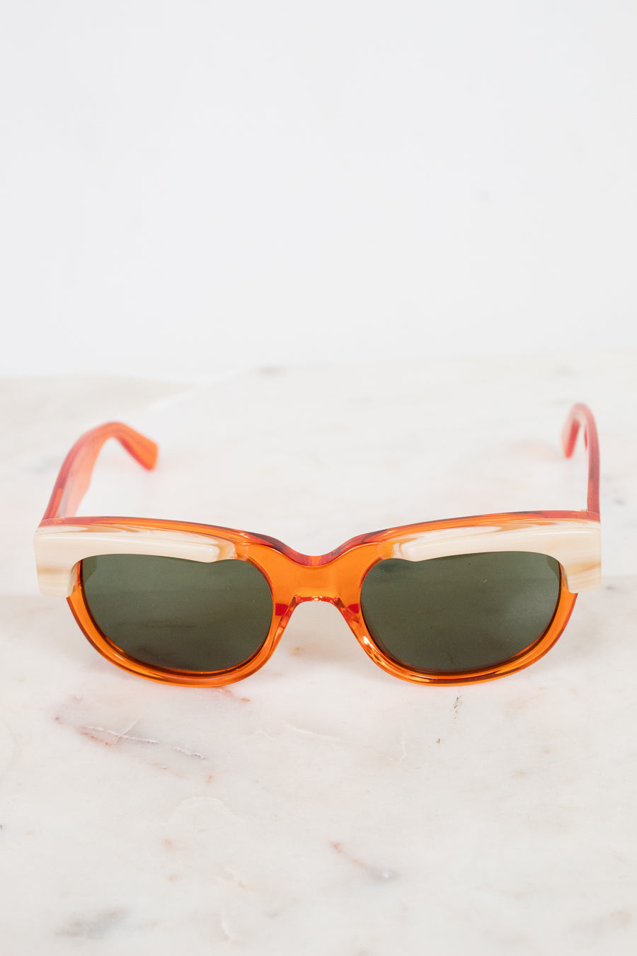 Orange acetate sunglasses with cream details and dark lenses resting on a white marble surface against a white background.