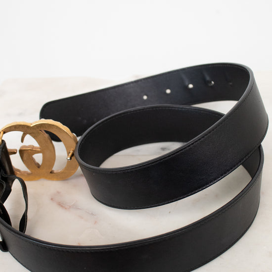 A black leather belt with a prominent gold-tone interlocking double-G logo buckle, coiled on a white marble surface against a white background.