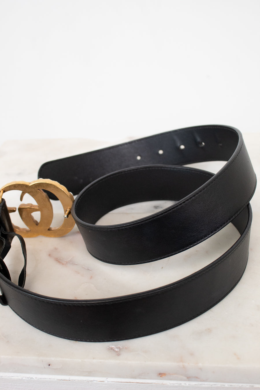 A black leather belt with a prominent gold-tone interlocking double-G logo buckle, coiled on a white marble surface against a white background.