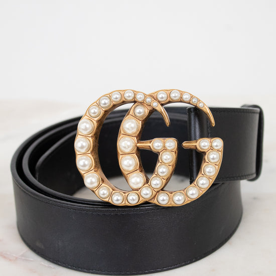 A black leather belt with a large interlocking G buckle embellished with gold-tone metal and white faux pearls, coiled on a light marble surface against a white background.