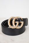 A black leather belt with a large interlocking G buckle embellished with gold-tone metal and white faux pearls, coiled on a light marble surface against a white background.