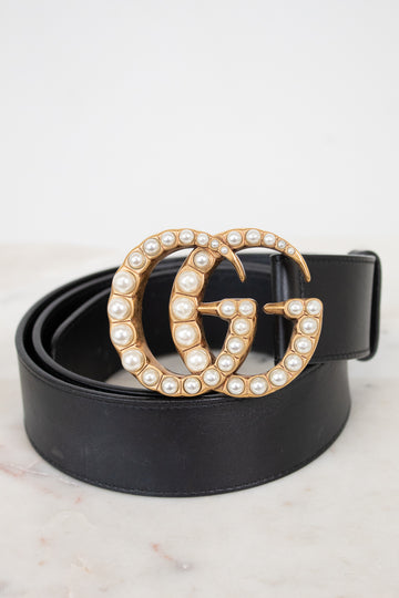 A black leather belt with a large interlocking G buckle embellished with gold-tone metal and white faux pearls, coiled on a light marble surface against a white background.