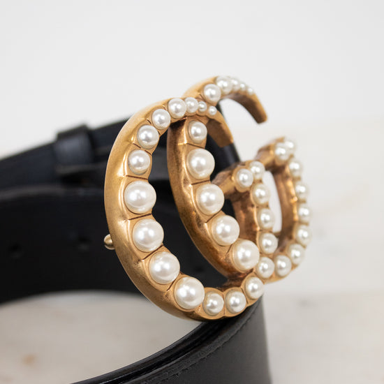 A close-up of a black leather belt with a large gold-tone double-G buckle embellished with white faux pearls, resting on a light surface.