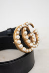 A close-up of a black leather belt with a large gold-tone double-G buckle embellished with white faux pearls, resting on a light surface.