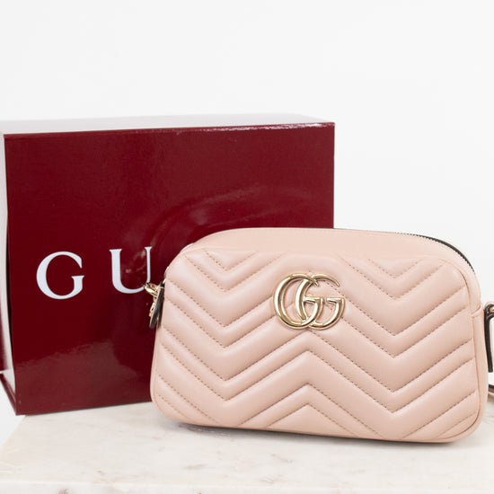 A light pink quilted leather camera bag with a gold interlocking G logo, shown next to its dark red Gucci box on a white marble surface.