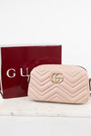 A light pink quilted leather camera bag with a gold interlocking G logo, shown next to its dark red Gucci box on a white marble surface.
