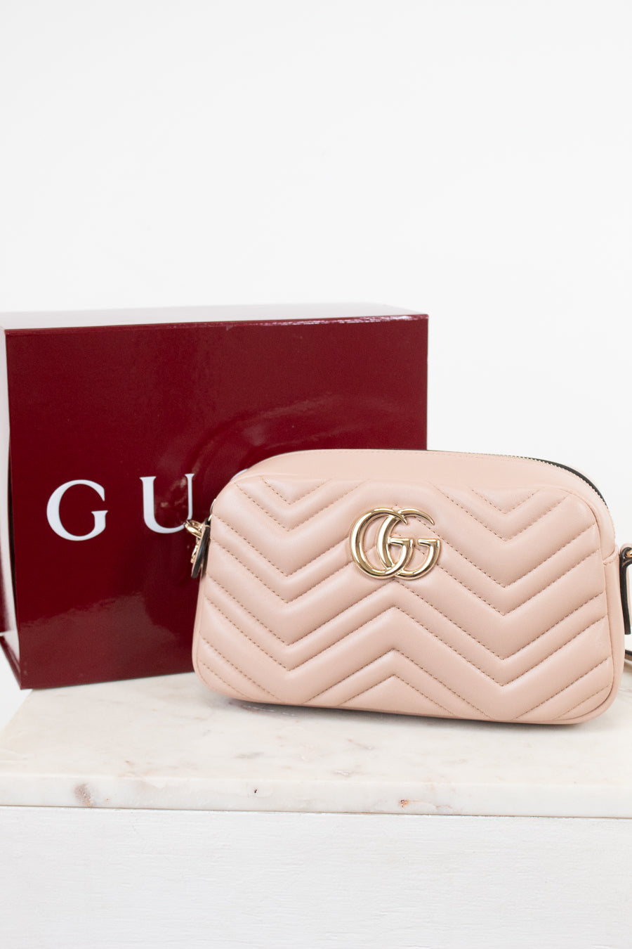 A light pink quilted leather camera bag with a gold interlocking G logo, shown next to its dark red Gucci box on a white marble surface.