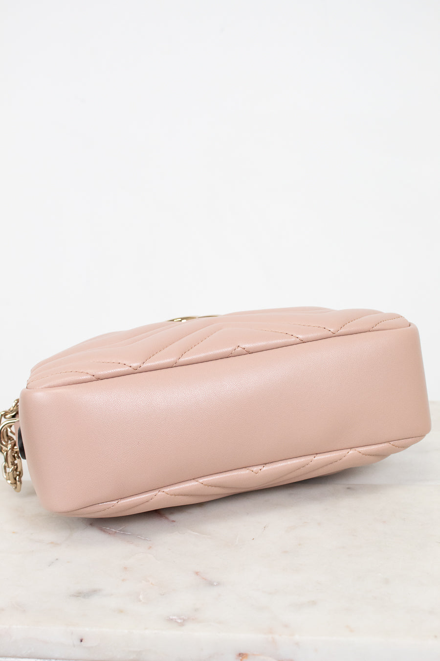 A light pink quilted leather shoulder bag shown from a side angle, resting on a white marble surface against a white background.