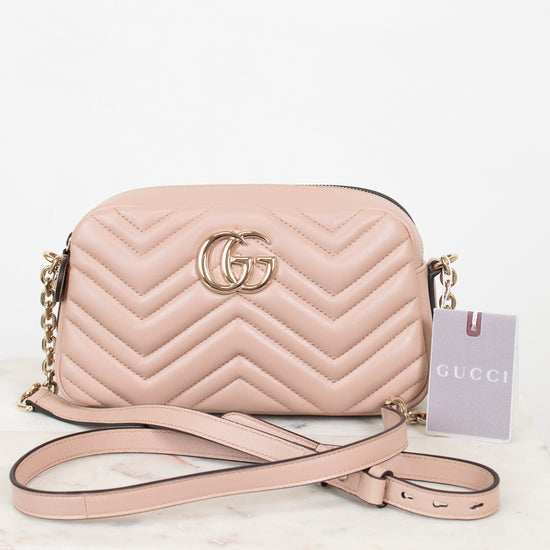 A light pink Gucci quilted leather crossbody bag with gold-tone hardware, shown on a white marble surface against a white background. The bag features the signature GG logo on the front, a chevron pattern, a gold chain strap, and a pink paper tag with the Gucci brand name.