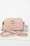 A light pink Gucci quilted leather crossbody bag with gold-tone hardware, shown on a white marble surface against a white background. The bag features the signature GG logo on the front, a chevron pattern, a gold chain strap, and a pink paper tag with the Gucci brand name.