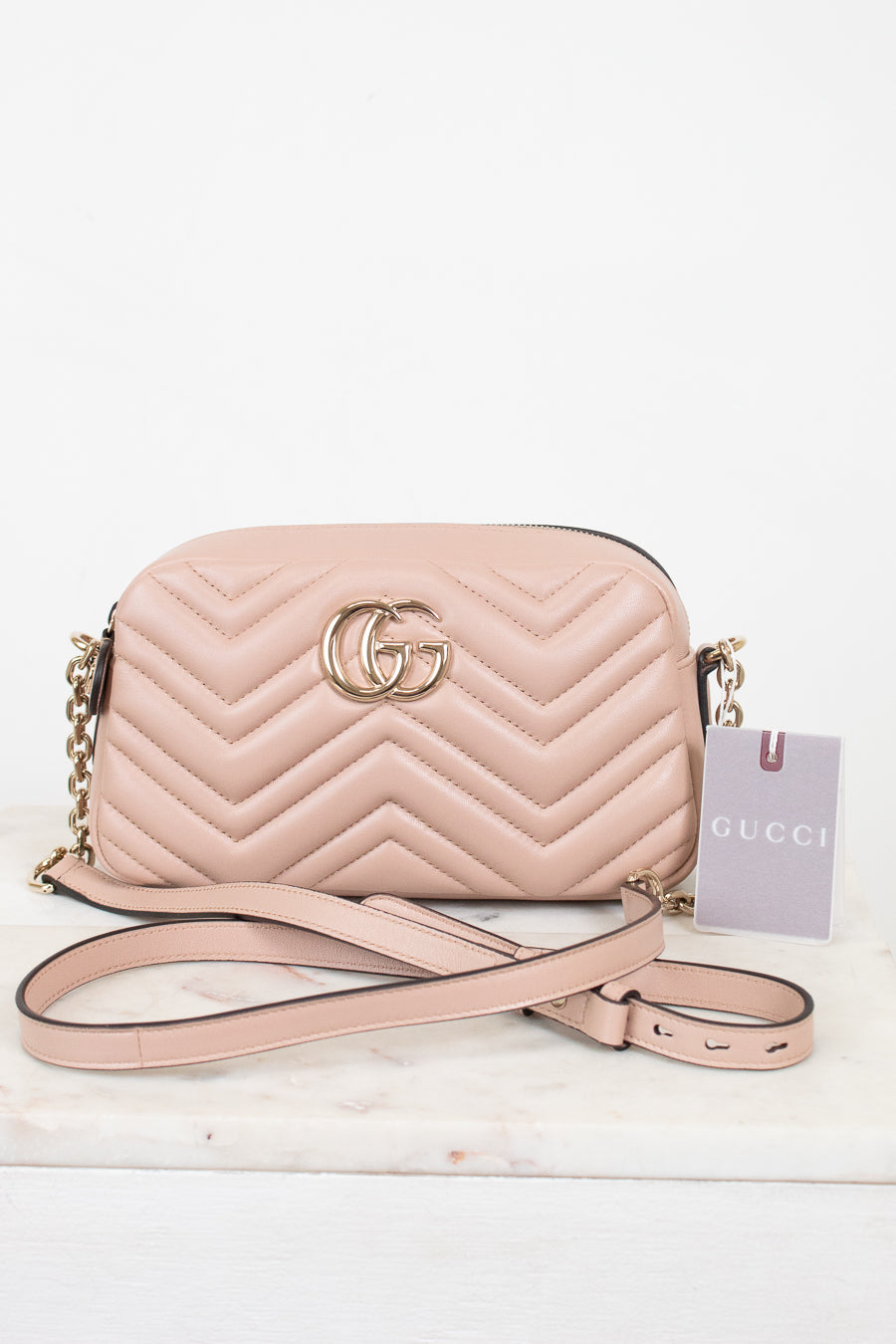 A light pink Gucci quilted leather crossbody bag with gold-tone hardware, shown on a white marble surface against a white background. The bag features the signature GG logo on the front, a chevron pattern, a gold chain strap, and a pink paper tag with the Gucci brand name.