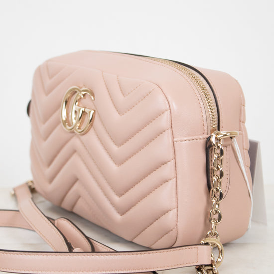 A light pink quilted leather camera bag with gold-tone hardware, featuring a chevron pattern, a gold interlocking G logo, and a matching shoulder strap on a white marble surface.