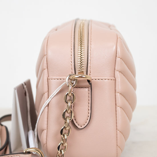 A close-up side view of a blush pink quilted leather crossbody bag with a gold-tone chain strap and zipper, resting on a light marble surface against a white background.