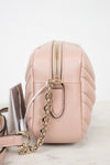 A close-up side view of a blush pink quilted leather crossbody bag with a gold-tone chain strap and zipper, resting on a light marble surface against a white background.