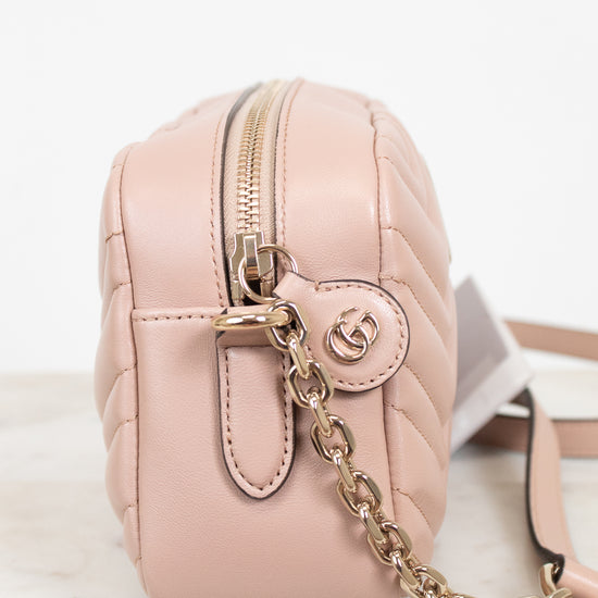 A light pink quilted leather crossbody bag with gold-tone hardware, featuring a front zip closure, a round interlocking GG logo charm, and a gold chain strap, displayed on a white marble surface against a white background.