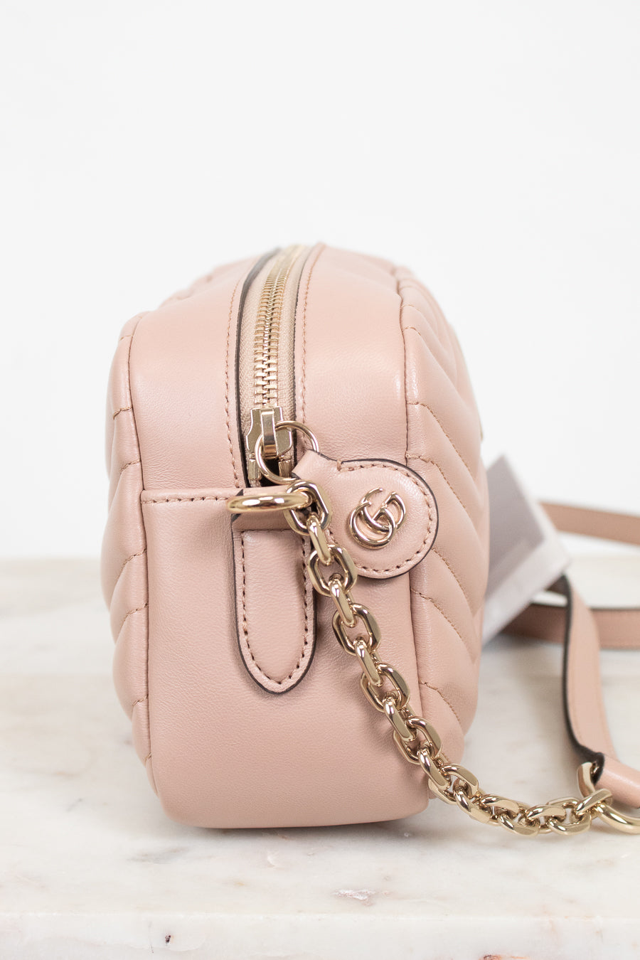 A light pink quilted leather crossbody bag with gold-tone hardware, featuring a front zip closure, a round interlocking GG logo charm, and a gold chain strap, displayed on a white marble surface against a white background.