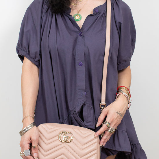 A woman wearing a dark purple short-sleeve button-up dress and holding a light pink quilted shoulder bag with a gold interlocking G logo, shown against a plain light gray background.