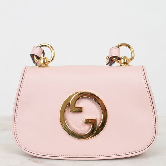 A light pink leather shoulder bag with a gold-tone interlocking G logo on the front and matching gold ring handles, displayed on a light marble surface against a white background.