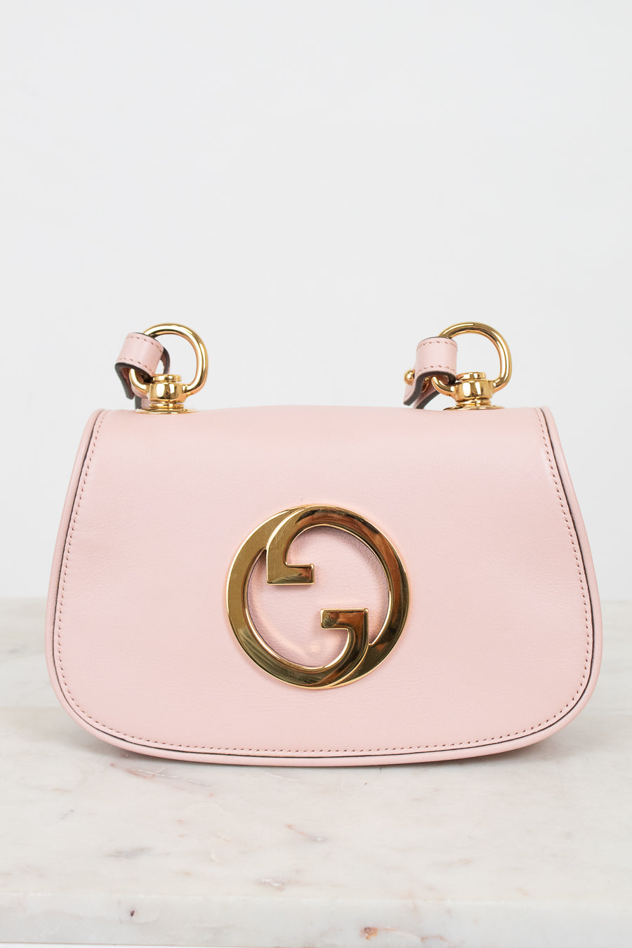 A light pink leather shoulder bag with a gold-tone interlocking G logo on the front and matching gold ring handles, displayed on a light marble surface against a white background.