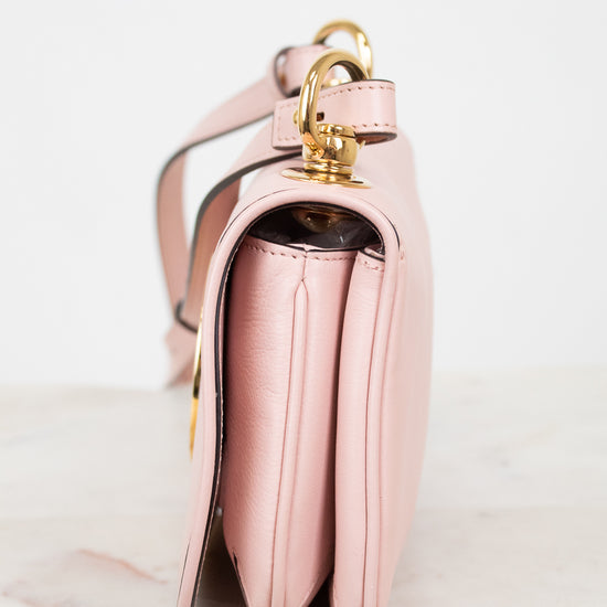 A close-up side view of a light pink leather handbag with gold-tone hardware, resting on a light marble surface against a white background.