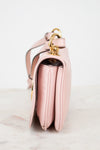 A close-up side view of a light pink leather handbag with gold-tone hardware, resting on a light marble surface against a white background.