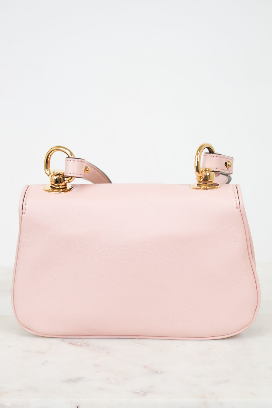 A light pink leather shoulder bag with gold-tone hardware and a matching pink strap, displayed on a light marble surface against a white background.