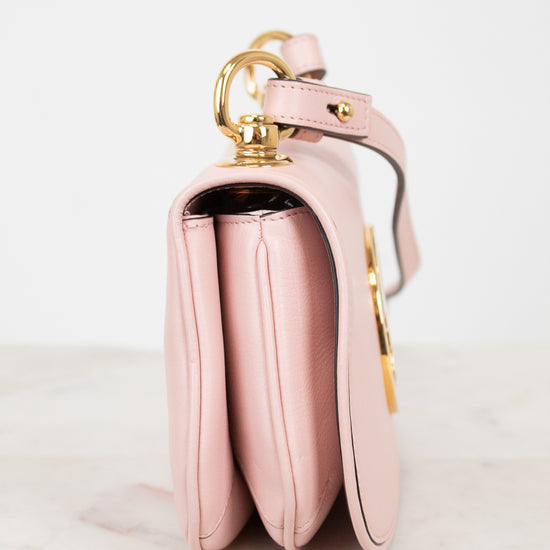 A light pink leather shoulder bag with gold-tone hardware, shown from a side profile view on a light marble surface against a white background.
