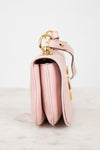 A light pink leather shoulder bag with gold-tone hardware, shown from a side profile view on a light marble surface against a white background.