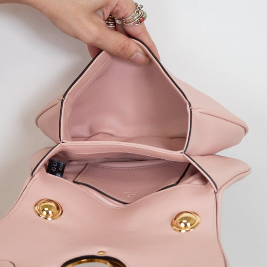 A close-up of a light pink leather handbag being opened by a person's hands, showing the interior pink lining, gold-tone hardware, and black piping details against a plain white background.