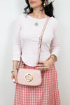 A woman wearing a light pink long-sleeve top and a red-and-white gingham skirt carries a light pink Gucci shoulder bag with a gold interlocking G logo. She is accessorized with gold jewelry against a plain white background.