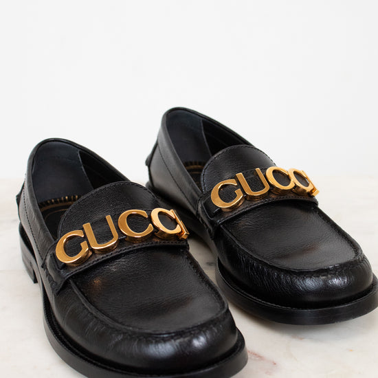 Black loafers with gold 'Gucci' logo on a light background