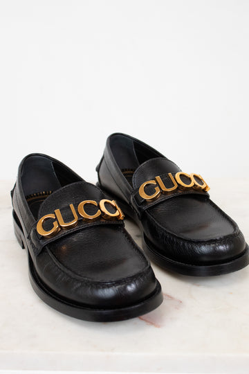 Black loafers with gold 'Gucci' logo on a light background